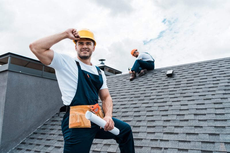 selective-focus-of-happy-repairman-holding-rolled-paper-while-coworker-repairing-roof